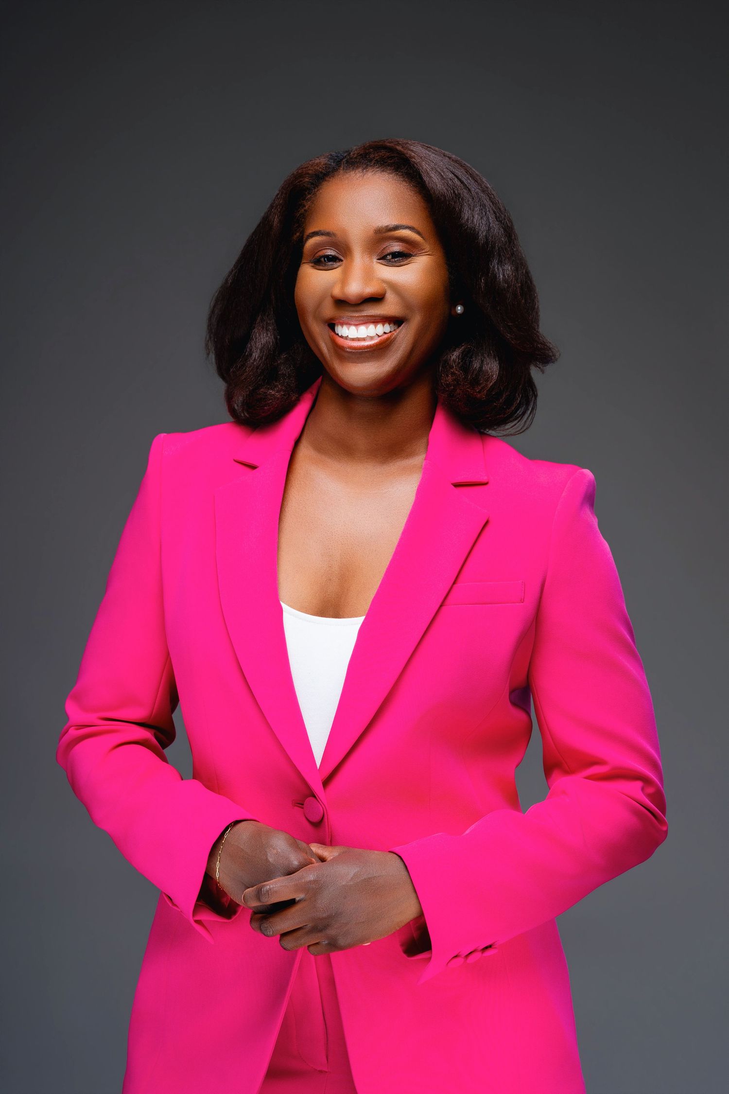 A confident Black woman in a bright pink blazer suit smiles warmly against a gray studio background.