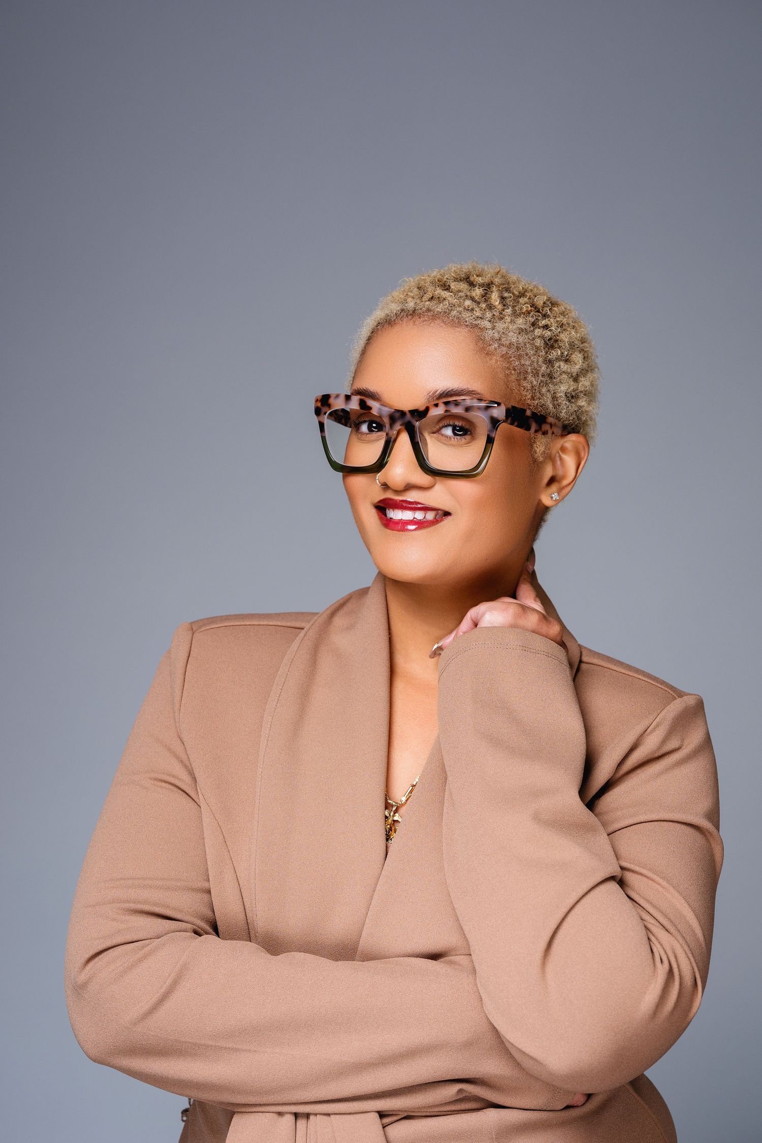 Professional woman with short blonde hair, tortoise glasses, and beige blazer posing confidently against gray background.