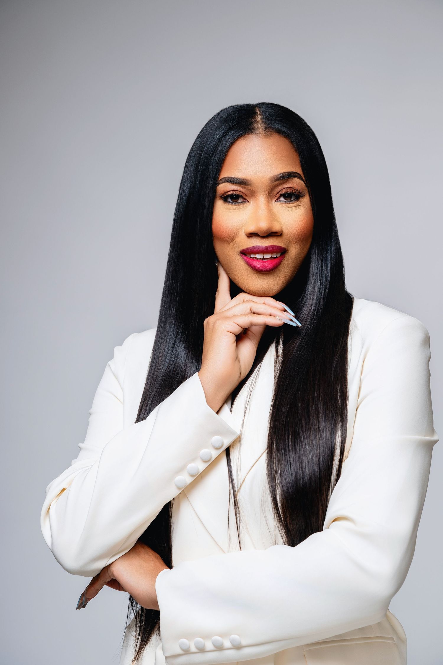 Professional portrait of a person in a crisp white blazer with long straight black hair and vibrant red lipstick against grey background.