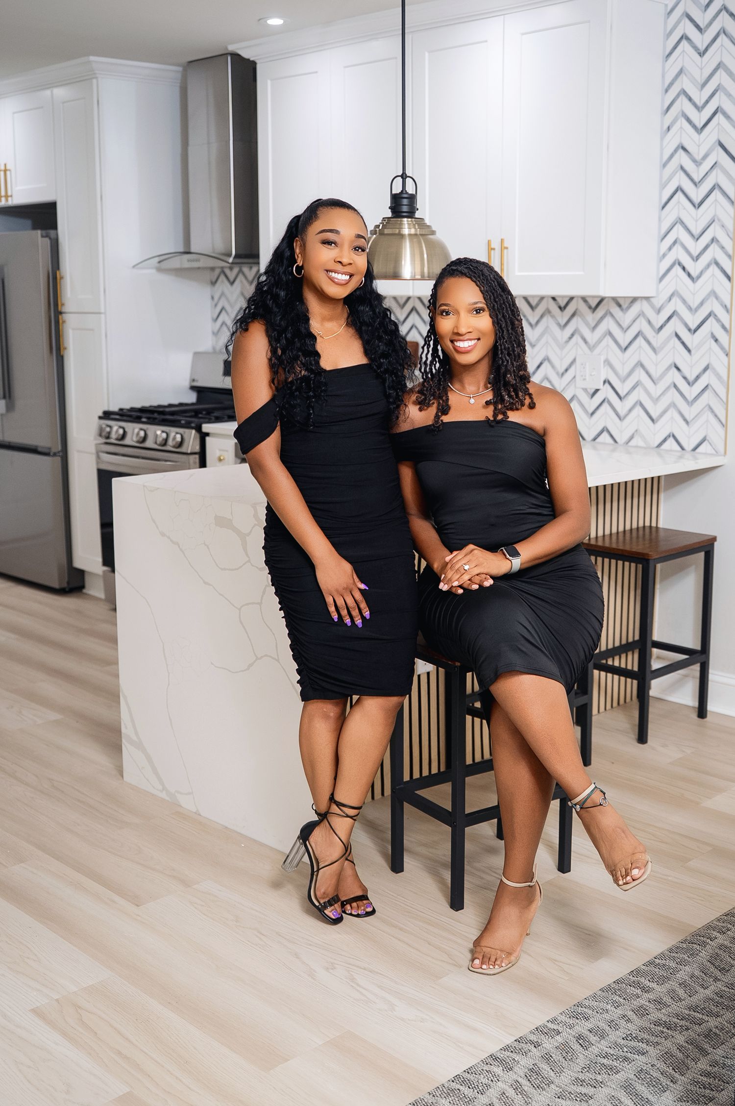 Two beautiful women wearing black outfits pose together on wooden flooring in a modern kitchen and living room setting.