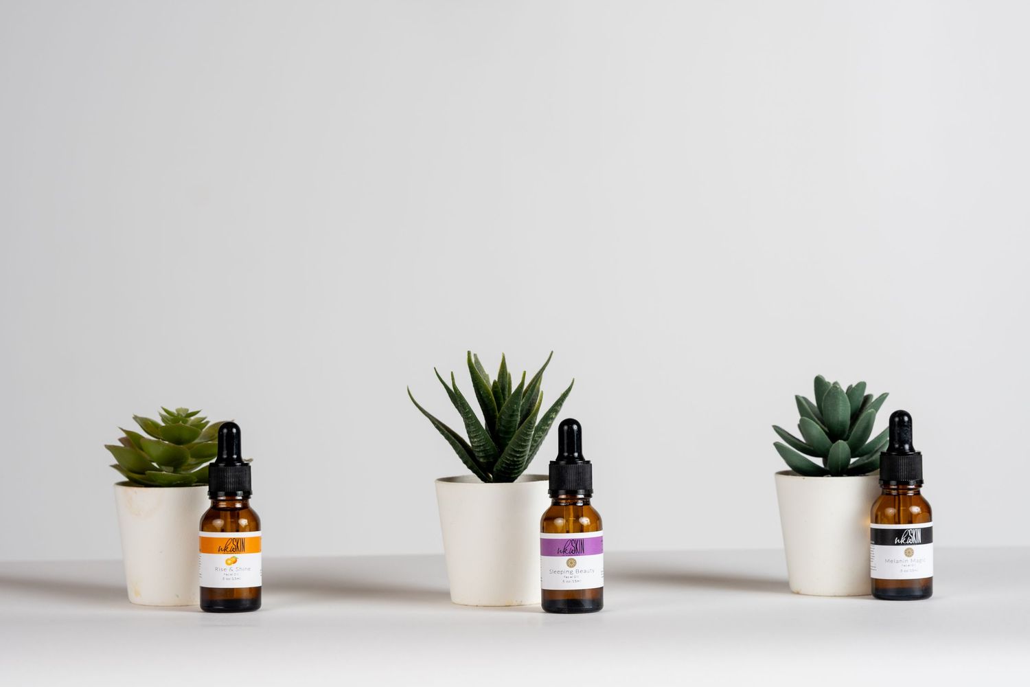 Three dropper bottles of essential oils displayed between small succulent plants in white pots on a white surface.
