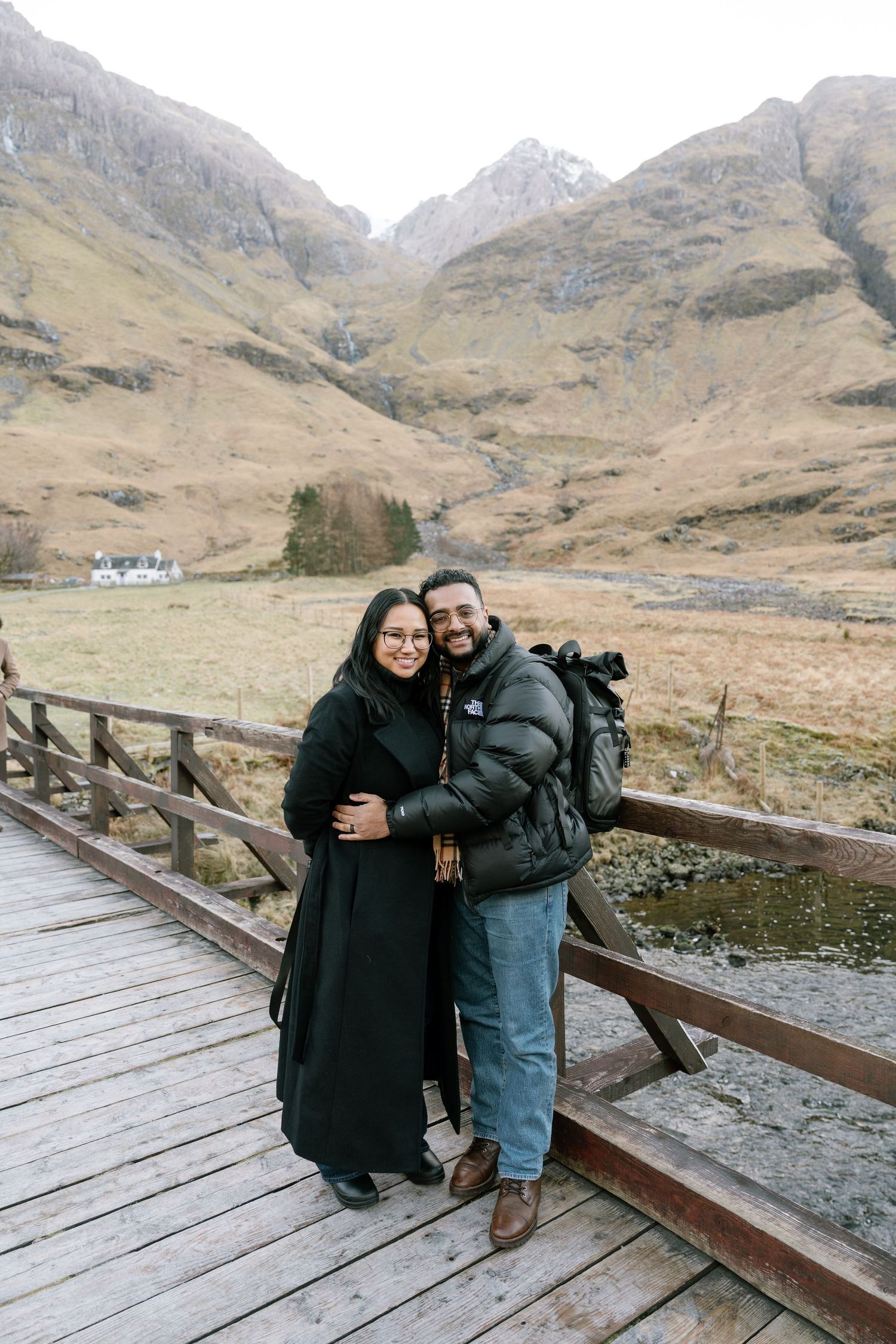 Couple embracing on wooden bridge in Glen Coe, Scotland, with dramatic mountain valley and snow-capped peaks behind them.