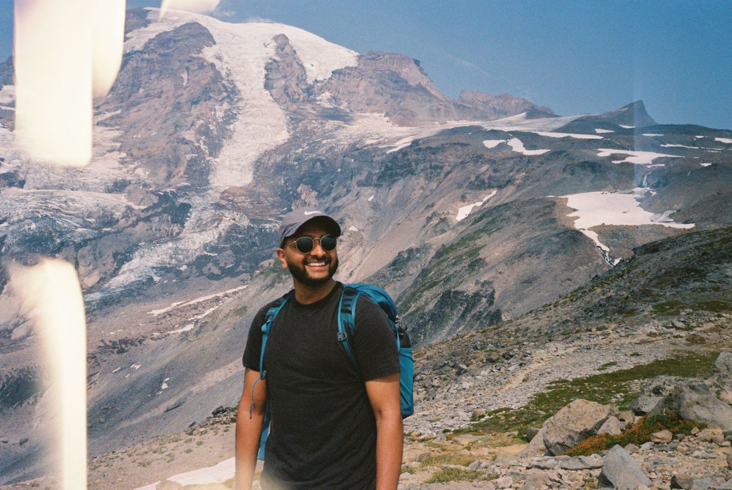 Smiling hiker in black shirt and cap poses before snow-capped mountain peaks on a clear sunny day.