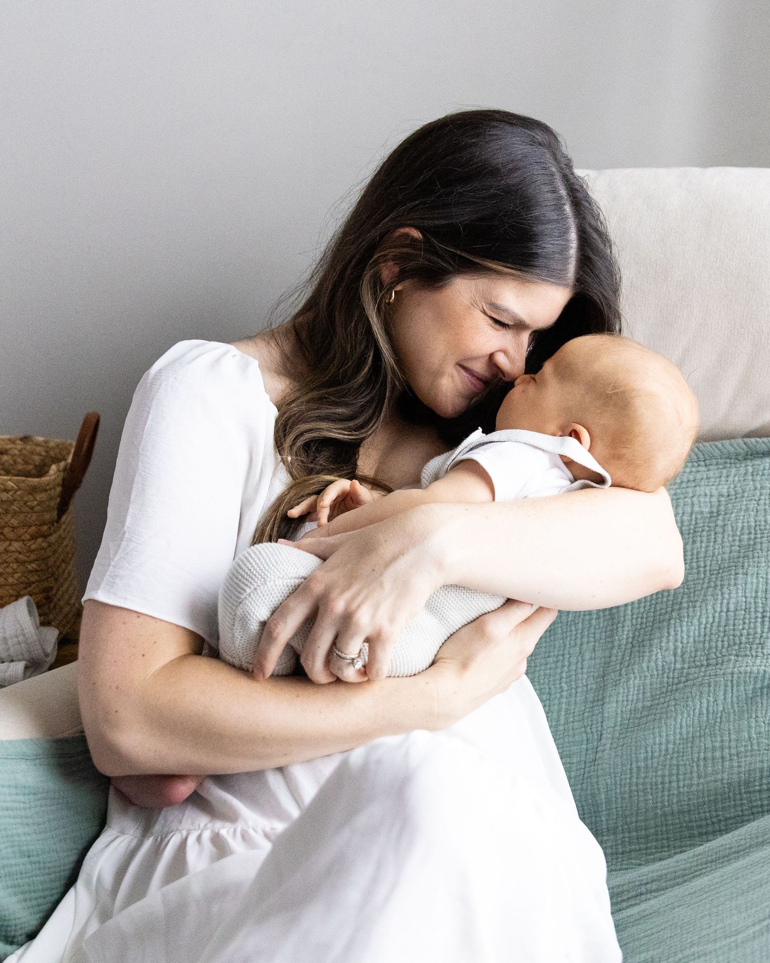 Photo of mother in white dress holding and touching noses with newborn son in white onesie