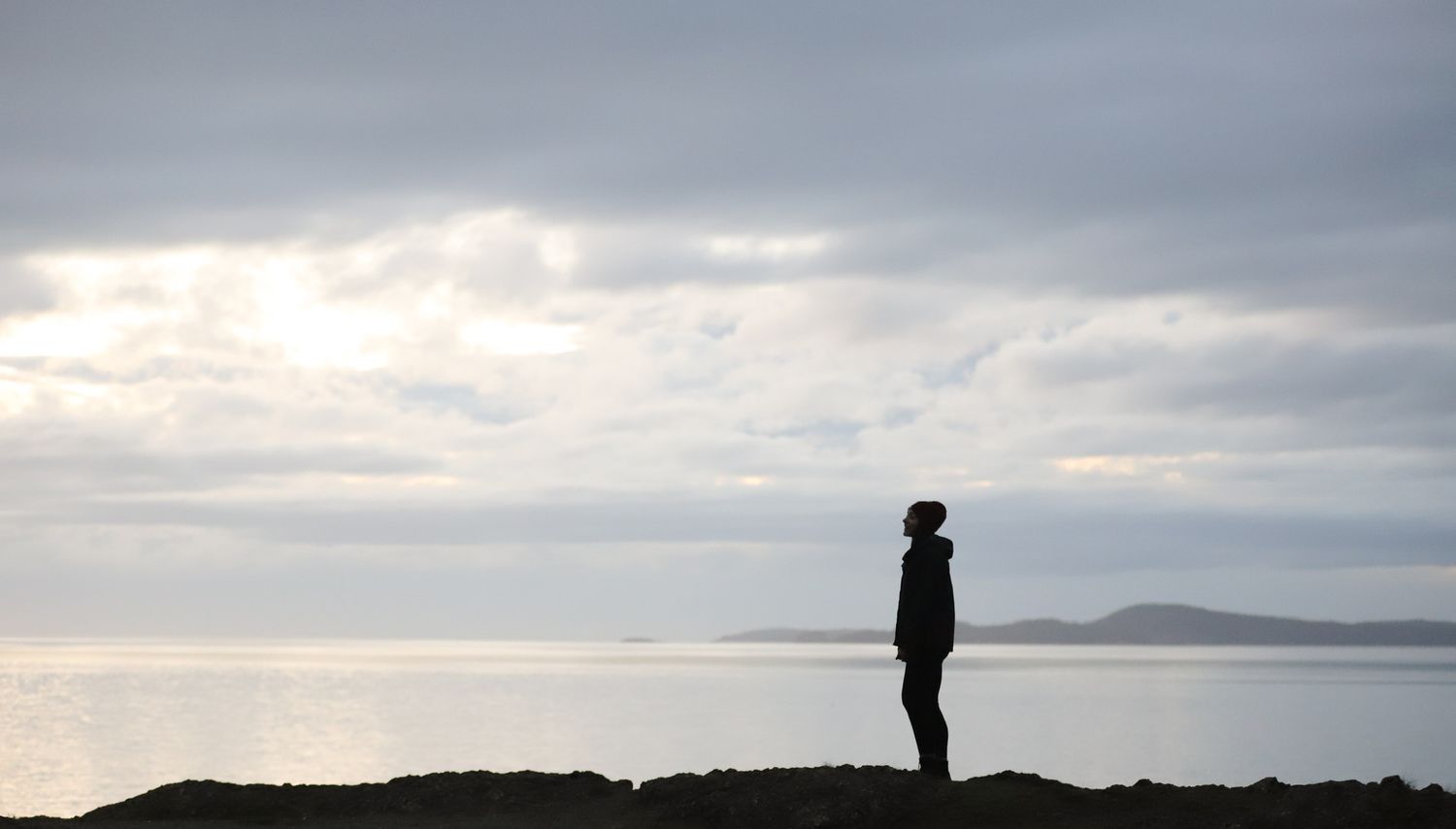 Silhouetted photo of woman standing on edge of mountain with water, mountains, and sky in the background
