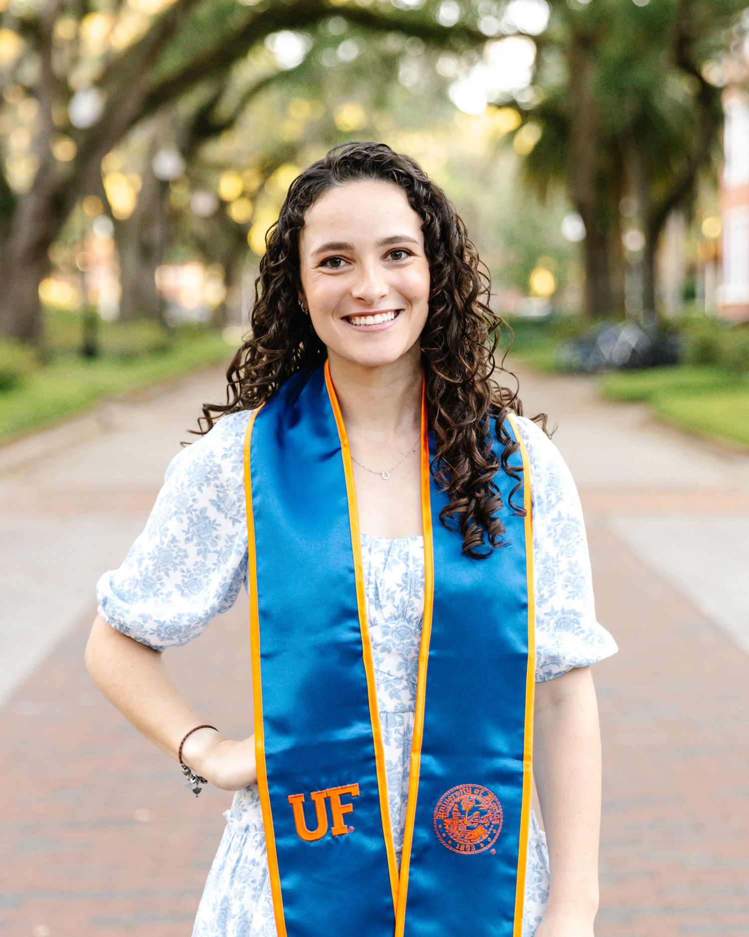 Photo of women with her hand on her hip, wearing a blue and white floral dress with a blue and orange graduation stole a
