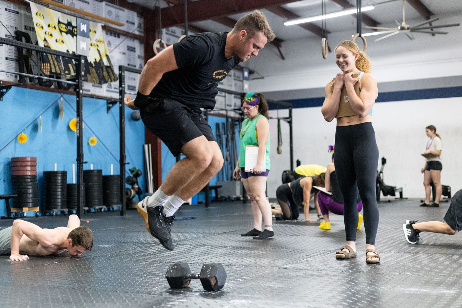 Photo of man jumping over dumbbell during a workout with a women holding a clipboard and smiling at him