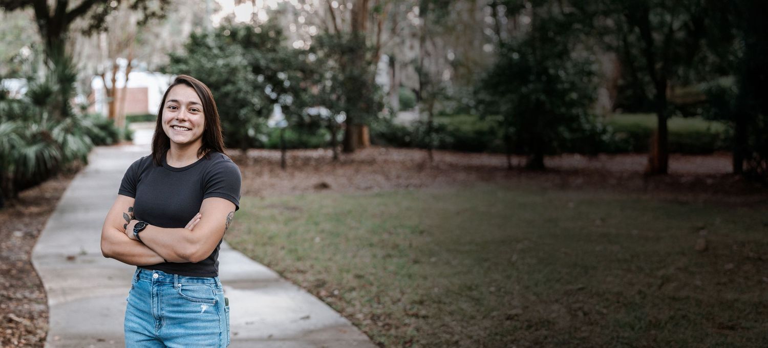 Photo of a woman wearing a black short sleeve shirt and jeans, standing with her arms crossed at a park