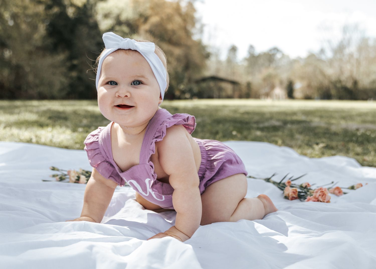 Photo of one year old baby crawling on white sheet with purple outfit and white bow headband on her head