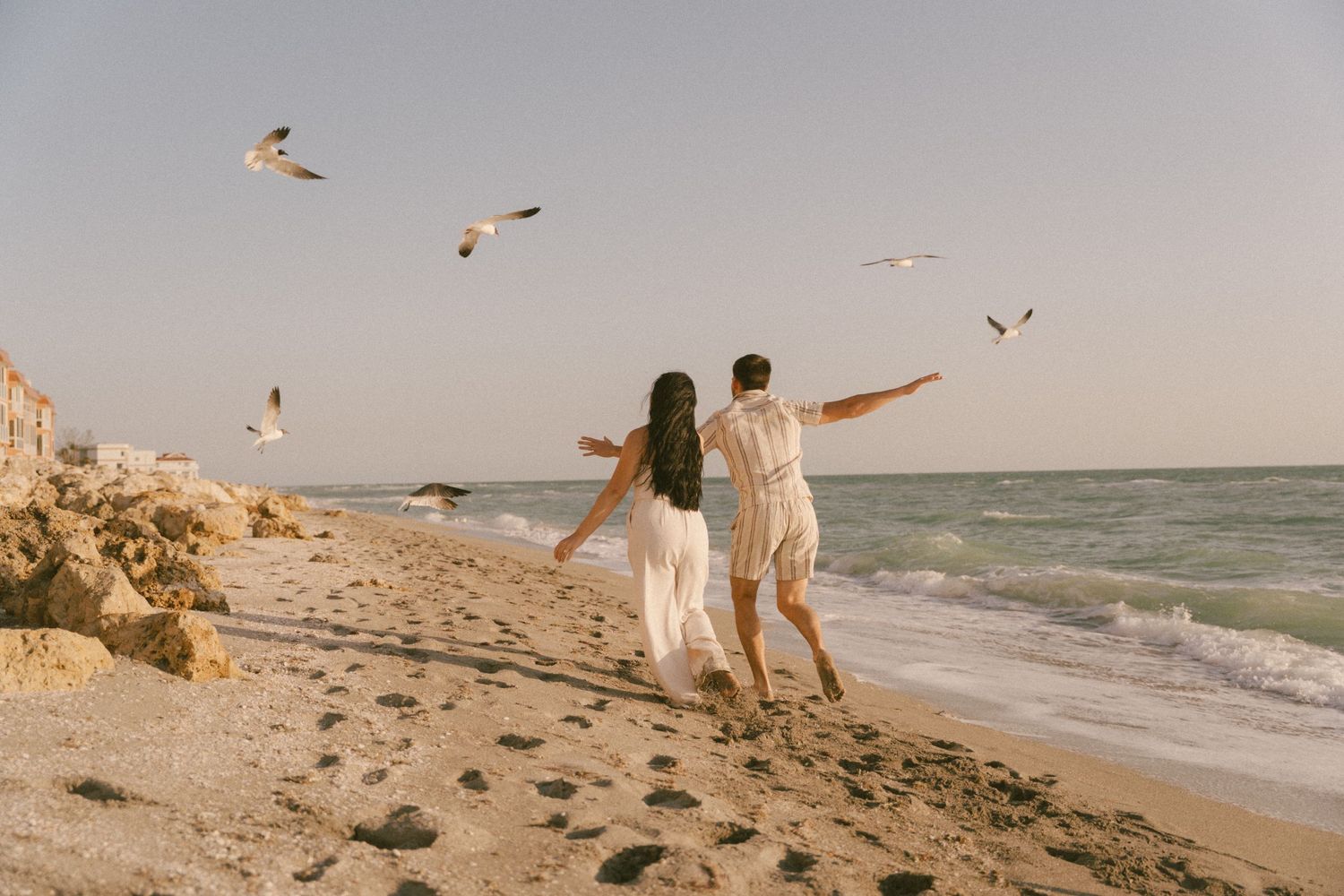 People walking on beach with seagulls flying overhead at sunset.