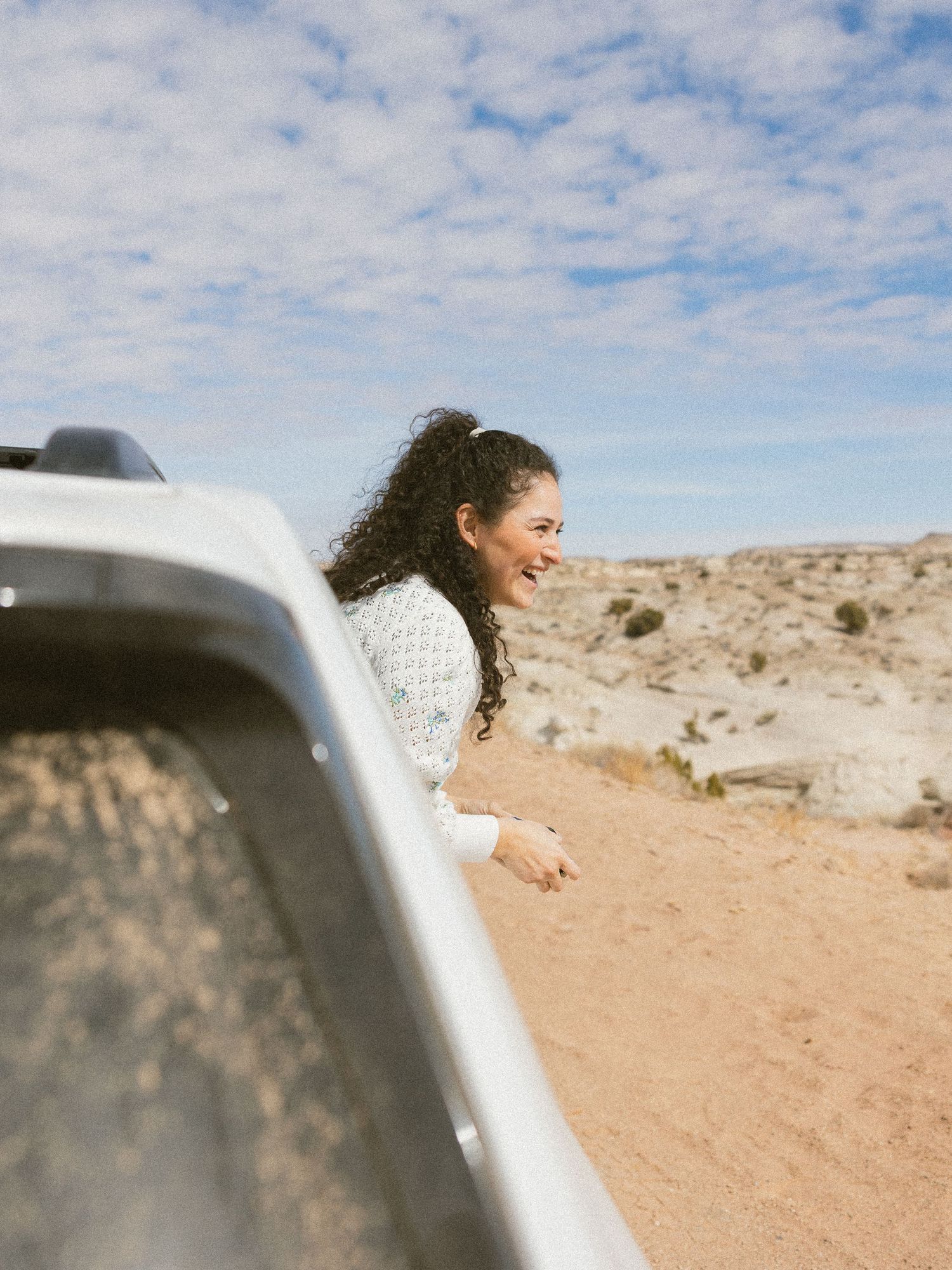 Person with curly hair leaning out car window against desert backdrop with cloudy sky.