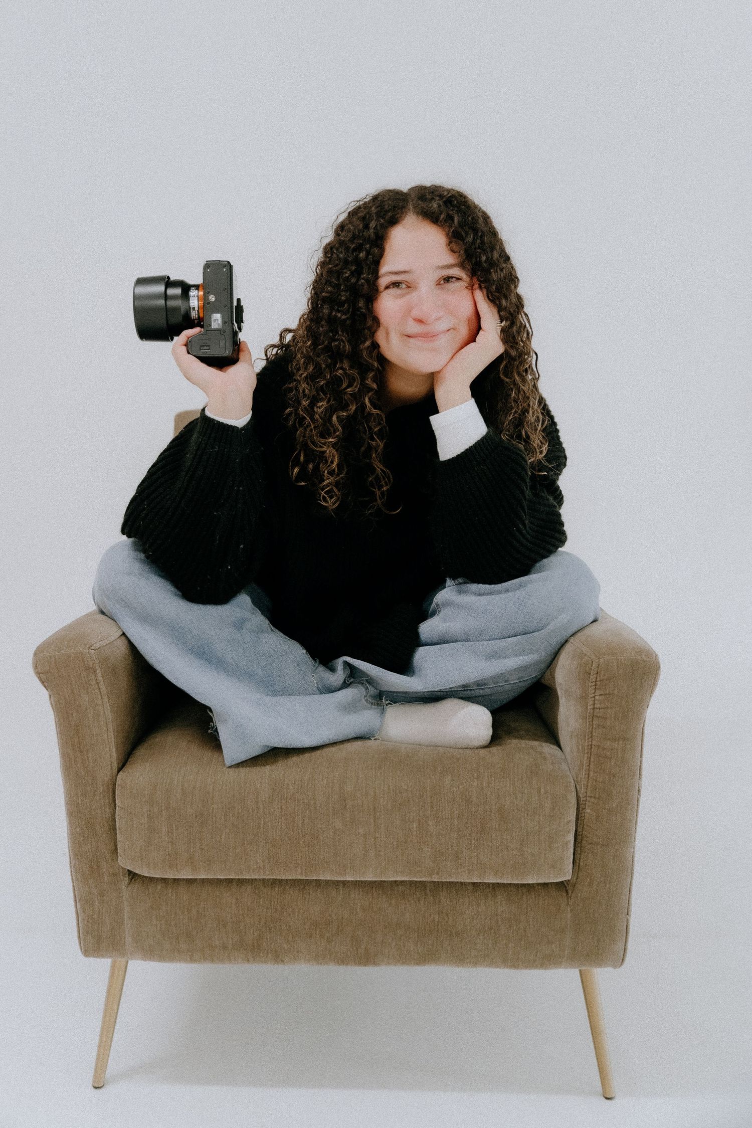 Person with curly hair lounging on tan couch with camera in minimalist studio setting.