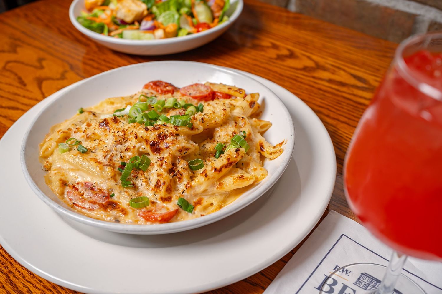 Plates of creamy pasta dishes with colorful garnishes served on a wooden restaurant table with red drinks.
