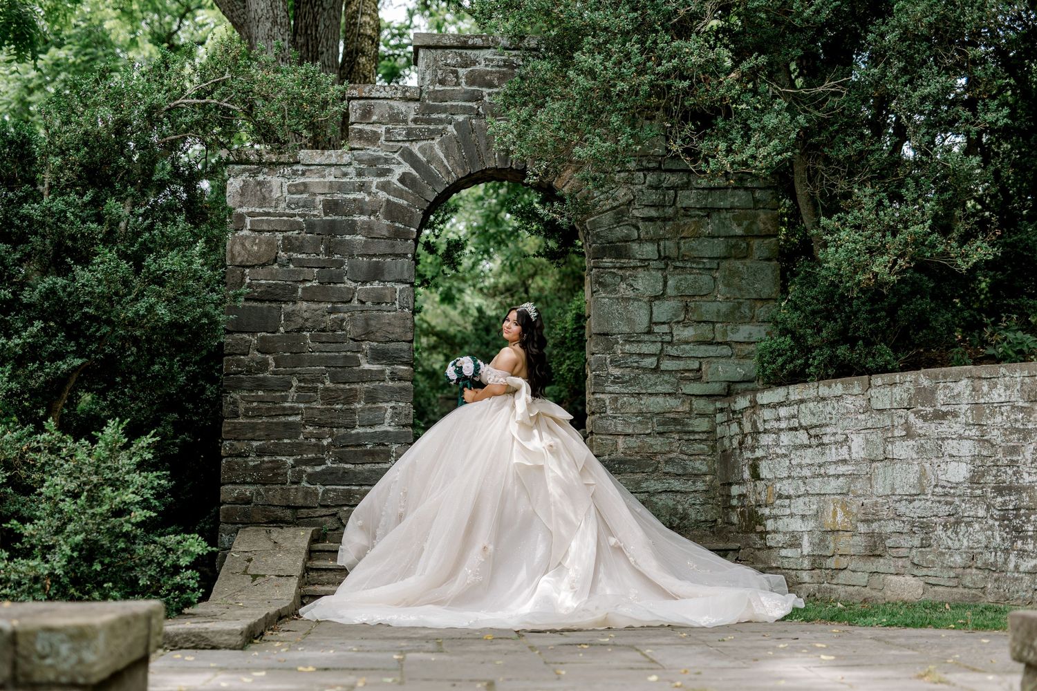 Bride in a voluminous ball gown poses before a stone arch gateway surrounded by lush greenery.