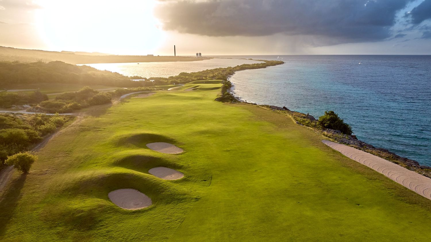 Dramatic sunset view of a coastal golf course with sand bunkers overlooking the ocean and a lighthouse in the distance.