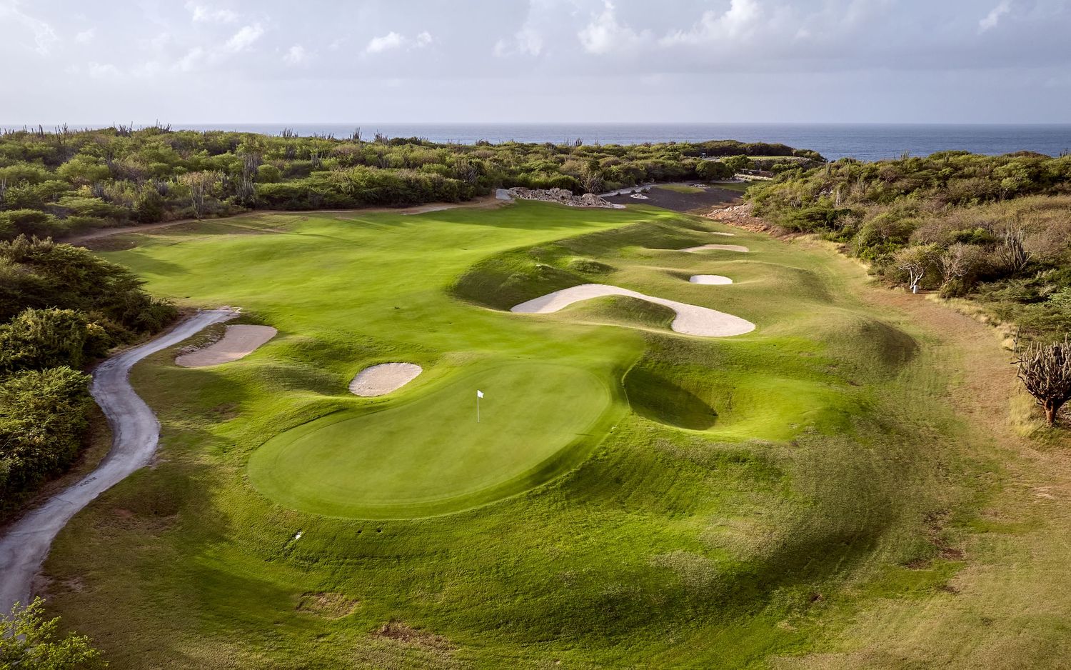 Aerial view of a coastal golf course with winding cart paths and white sand bunkers surrounded by natural vegetation.