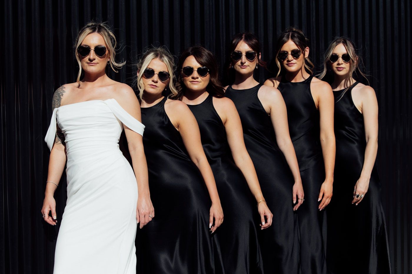 A group of people wearing black dresses and sunglasses stand in a row next to someone in a white dress.