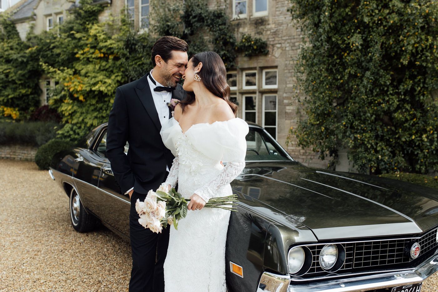 A couple in wedding attire embraces beside a classic black vintage car during an outdoor photo session.