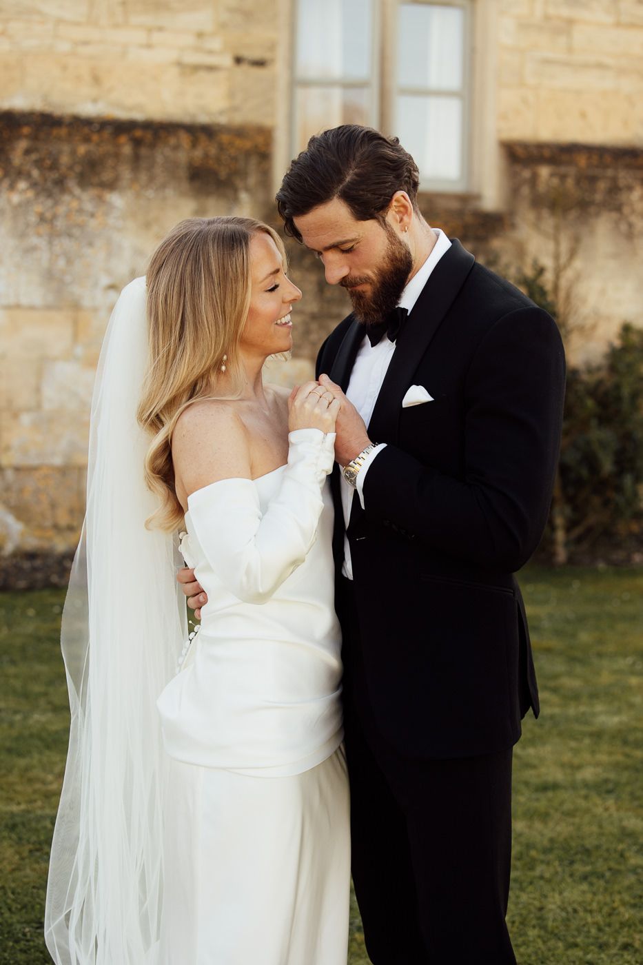 A romantic wedding photo shows a couple in classic attire sharing an intimate moment outdoors against a stone wall.