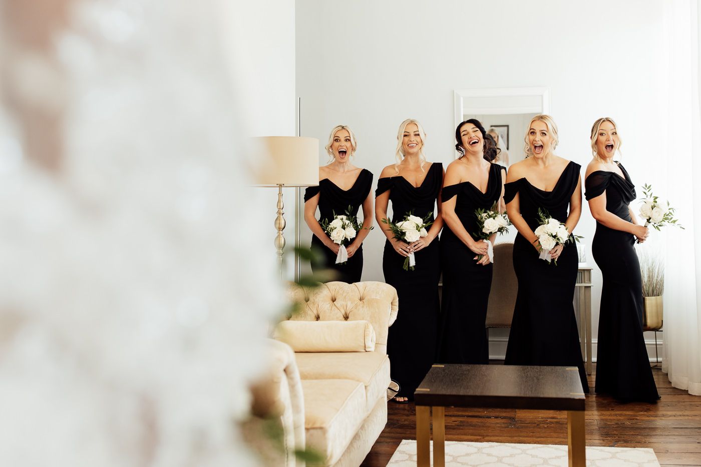 Bridesmaids in elegant black dresses stand together holding white bouquets in a bright interior space at Syrencot