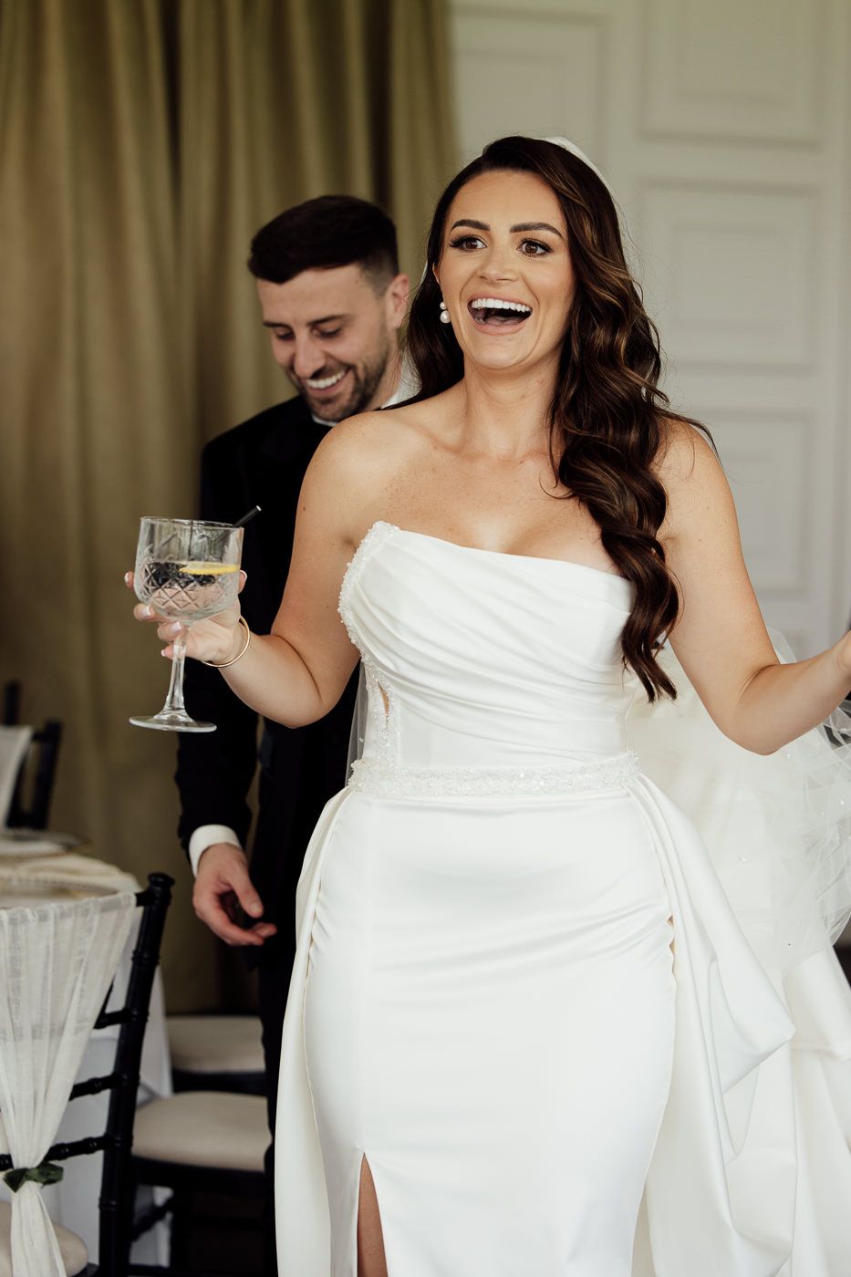 Joyful bride in elegant white strapless gown holding wine glass and laughing during wedding celebration.
