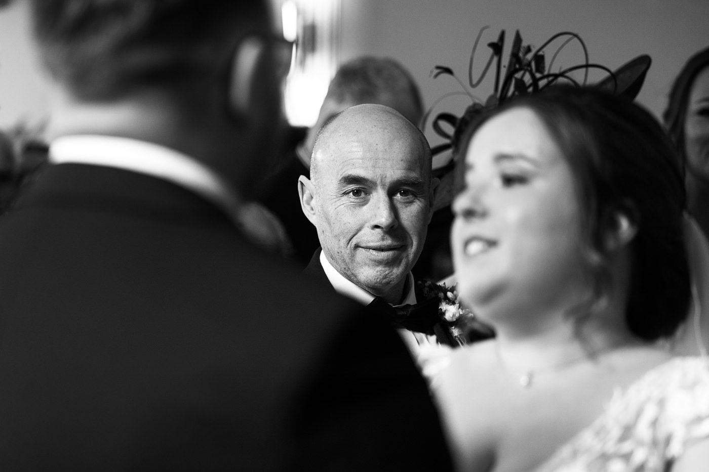 A black and white photograph captures a serious figure amid a wedding ceremony looking directly at the camera.