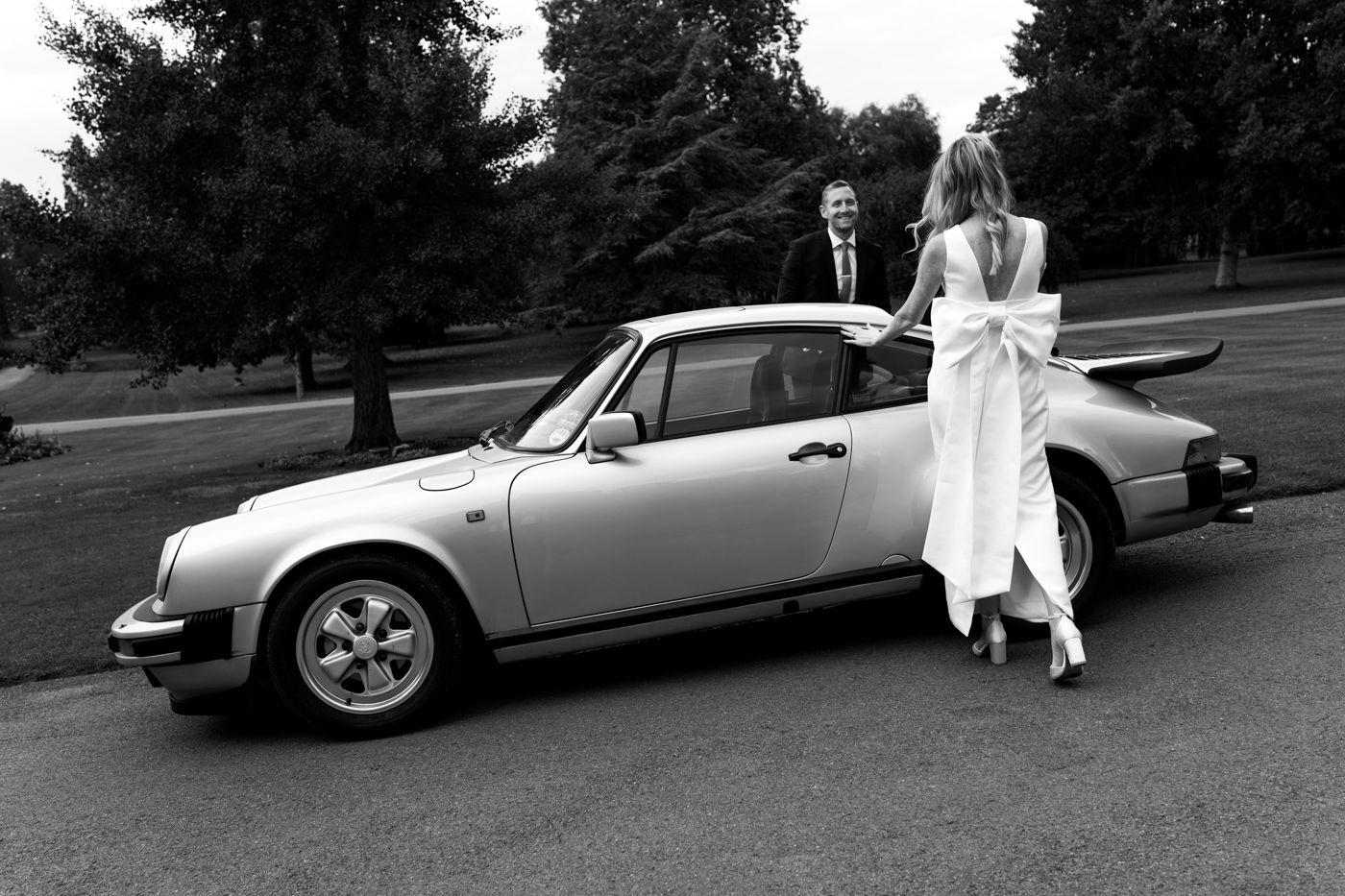 Classic Porsche 911 parked on a road with a couple in formal attire standing beside it in black and white photography.