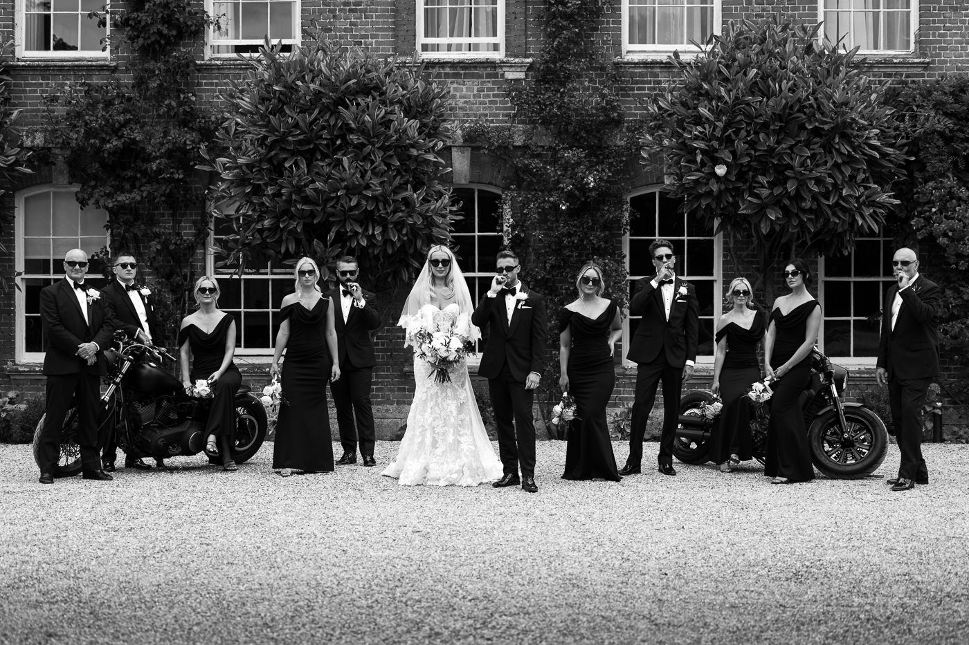 A black and white wedding portrait of a bridal party lined up on a gravel path in front of a brick building. Mob style