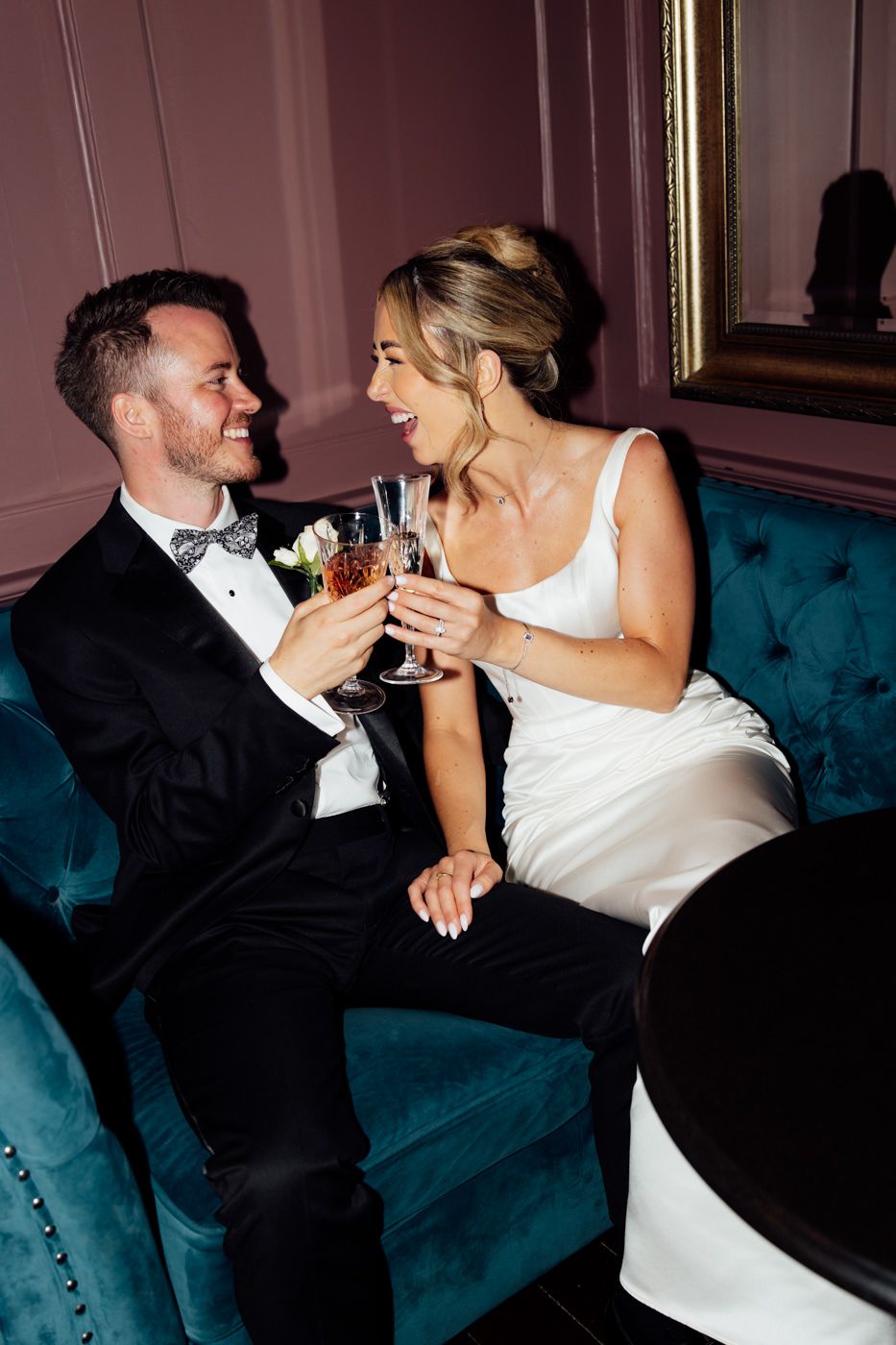 A couple in wedding attire shares a romantic moment on a teal velvet couch during their celebration.