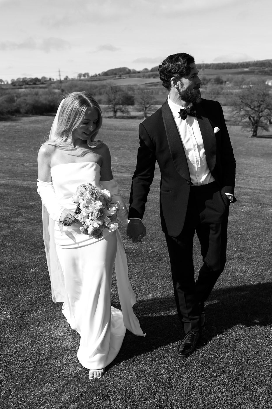 A black and white wedding photo showing a couple outdoors on a grassy hill, with the bride holding a floral bouquet.