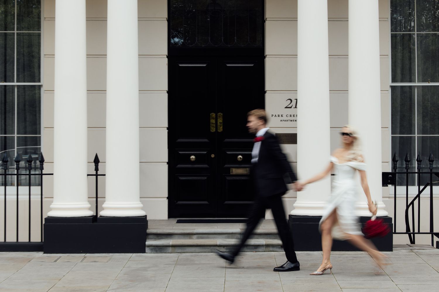 Two people hurriedly walk past a white townhouse entrance with black door and elegant columns in London.