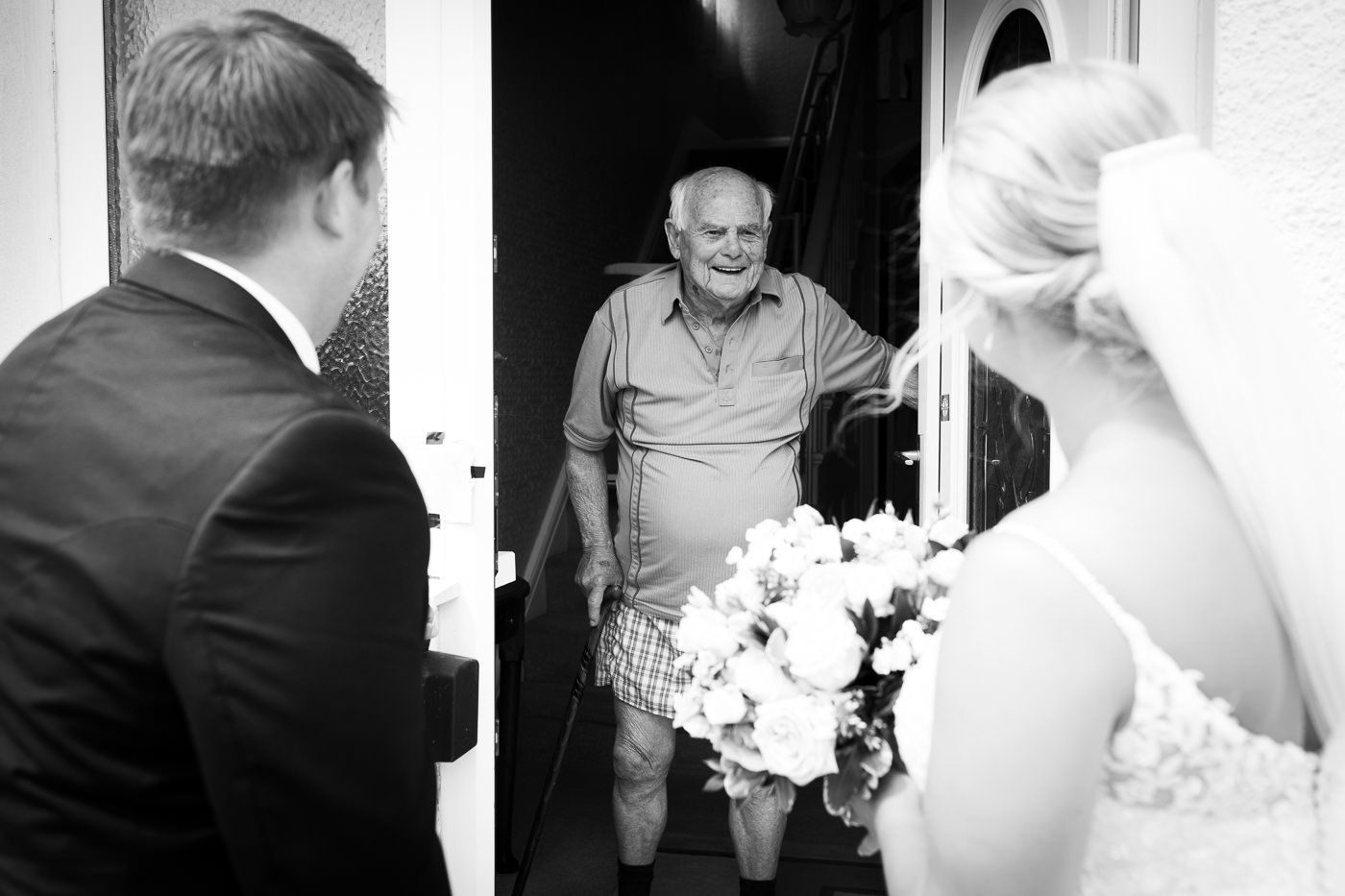 A candid wedding photo of a bride and groom visiting the bride's grandad who is standing at the door in his pants.