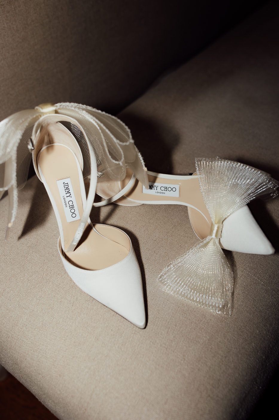 Elegant white Jimmy Choo bridal heels with strappy design and delicate mesh bow detail on beige surface.
