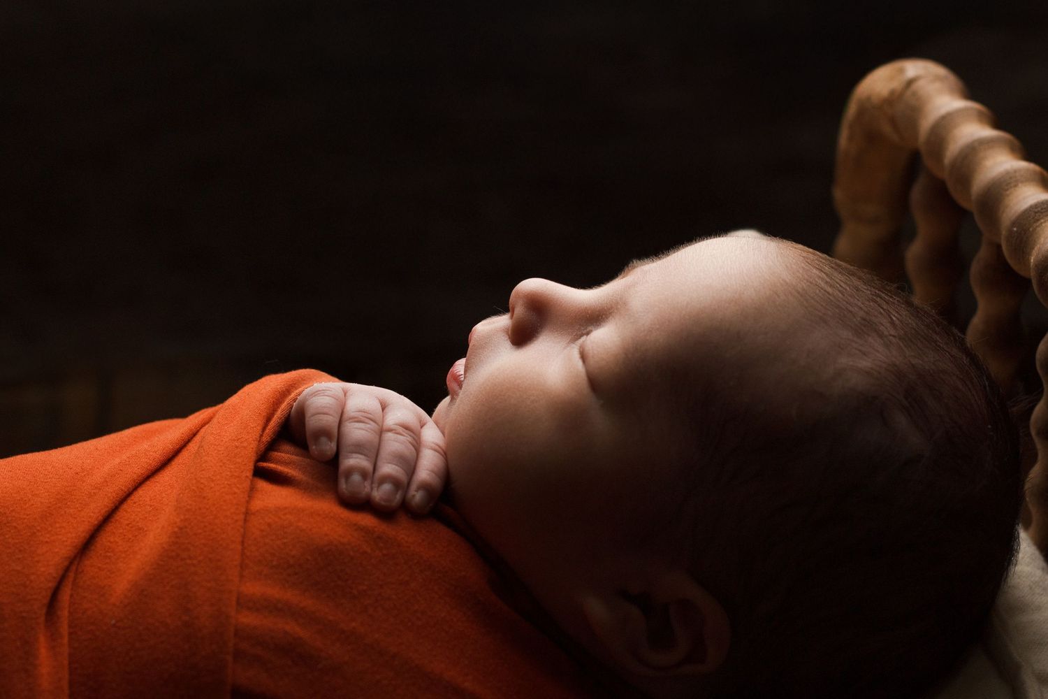 Newborn baby sleeping in a small wooden bed wrapped in a warm rust-colored wrap side profile during a styled newborn session.