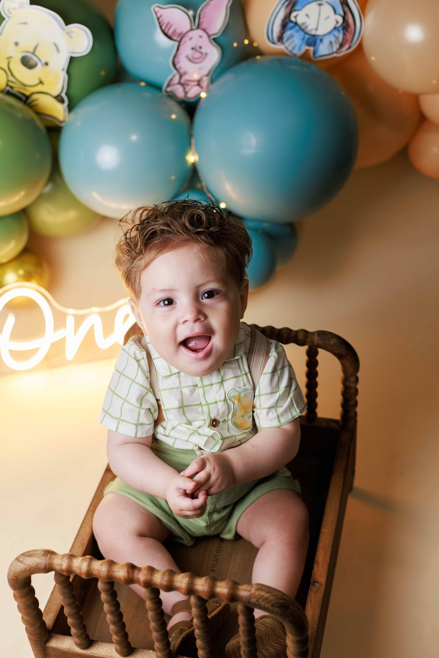 Baby boy  wearing a cream and green suspenders sitting on a little bed surrounded by blue and green balloons during a Cake Smash Session