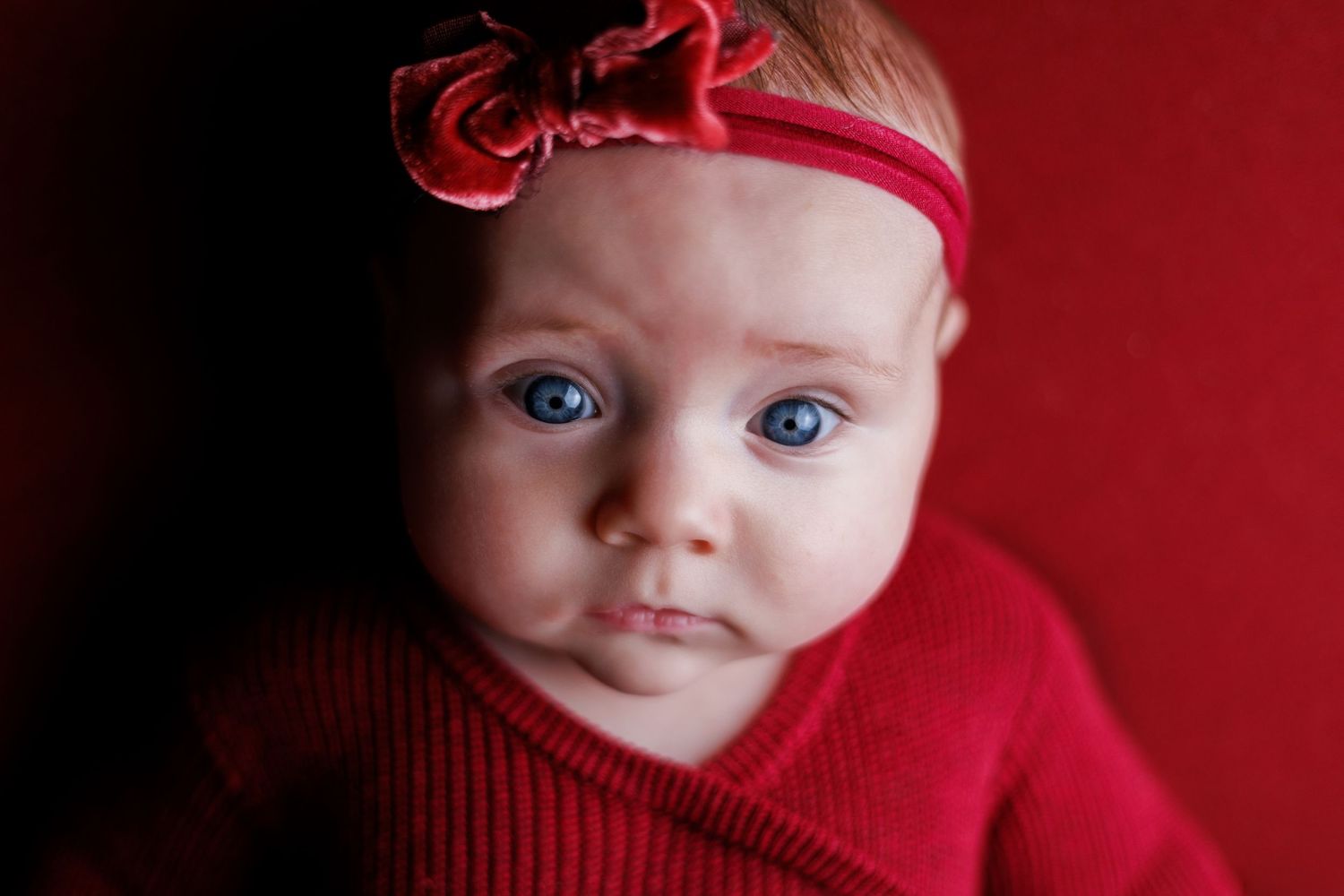 Adorable baby girl with striking blue eyes wearing a red outfit and floral headband against a red background.