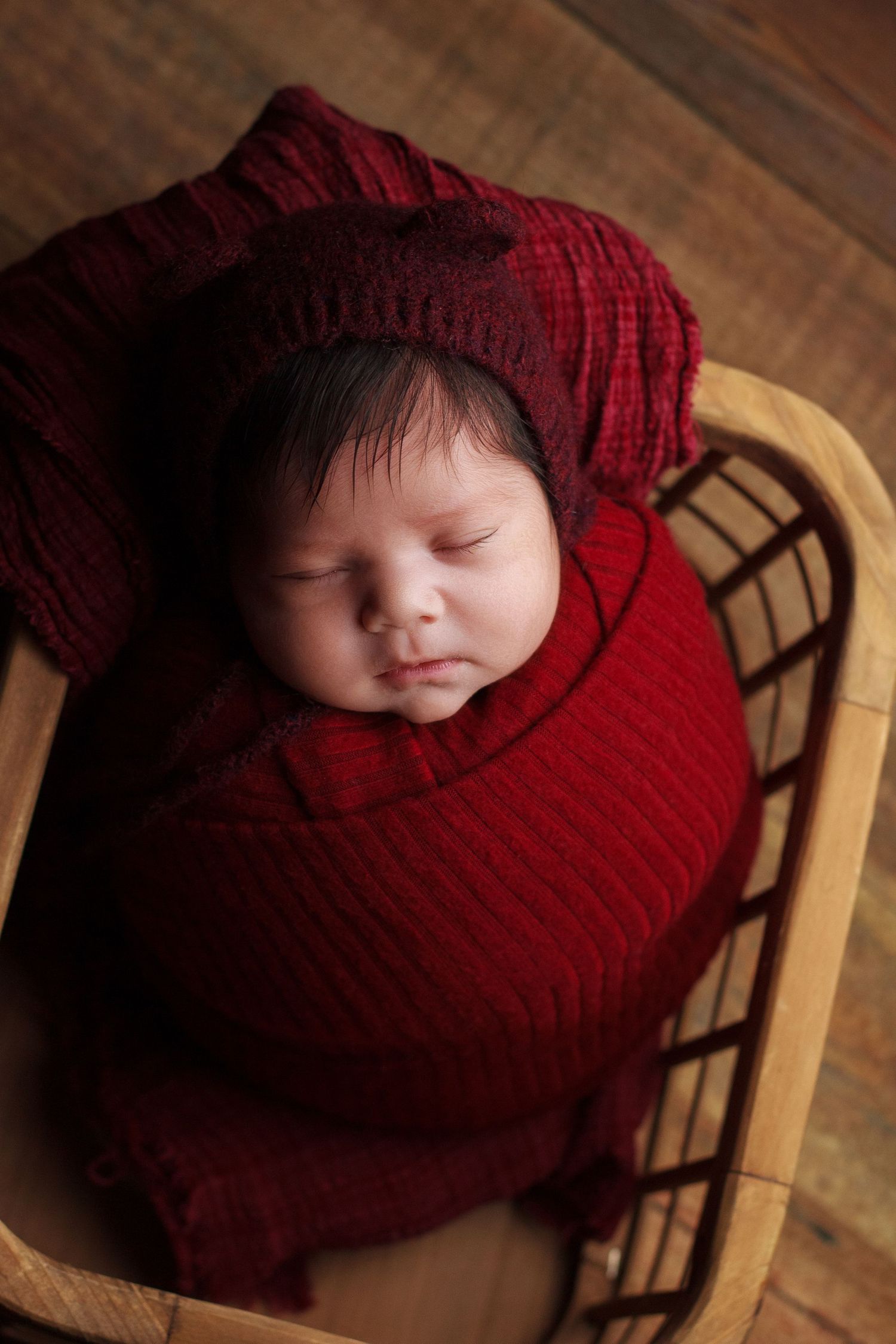 Newborn baby girl wrapped in deep red knit resting in a wooden basket during a professional newborn photography session in Dalton, Georgia.