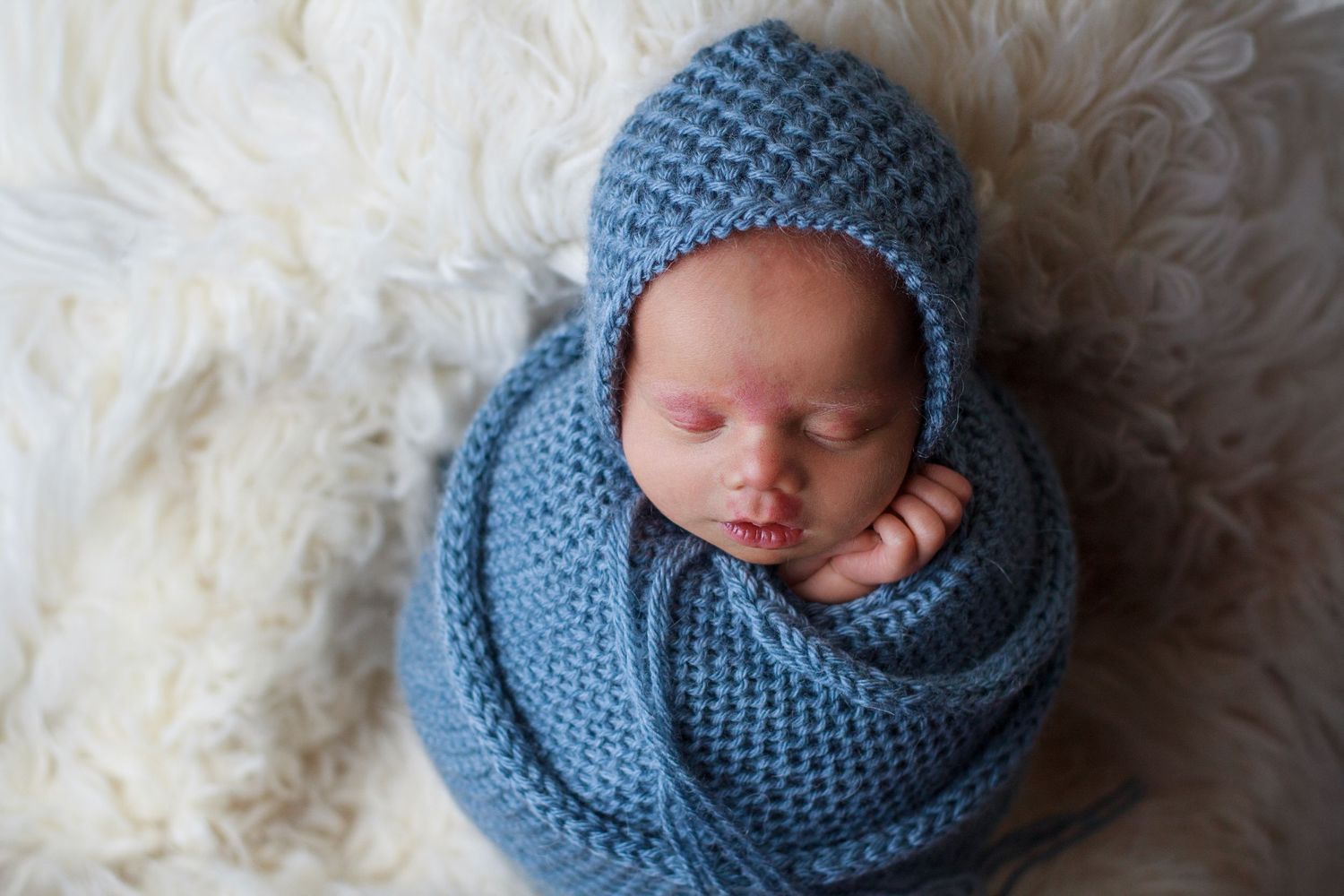 Newborn baby boy wrapped in a blue knit wrap with matching blue knit bonnet resting on a Flokati during a Dalton Georgia Newborn Session
