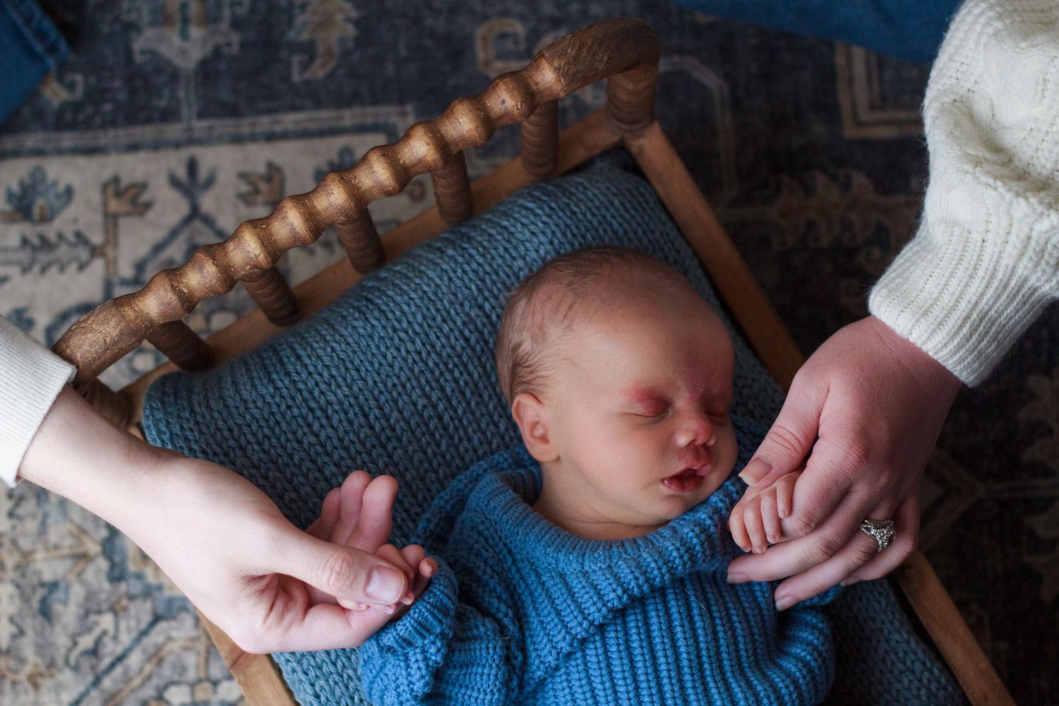 Newborn baby boy lying in a small wooden bed while parents gently hold his hands during a newborn session.