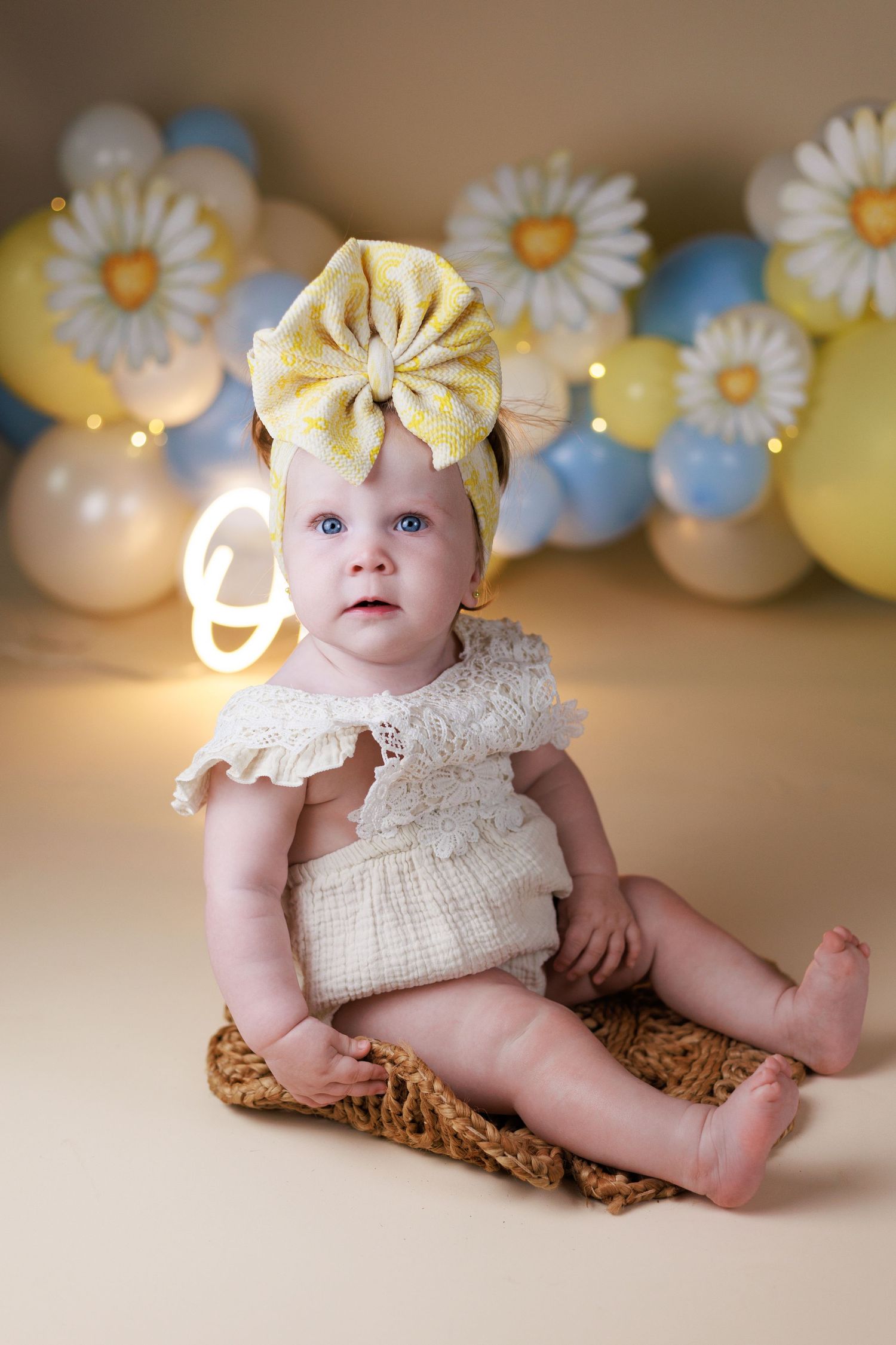 Baby girl dressed in a cream lace romper sits in front of a pastel yellow and blue balloon background with daisies for a Cake Smash session