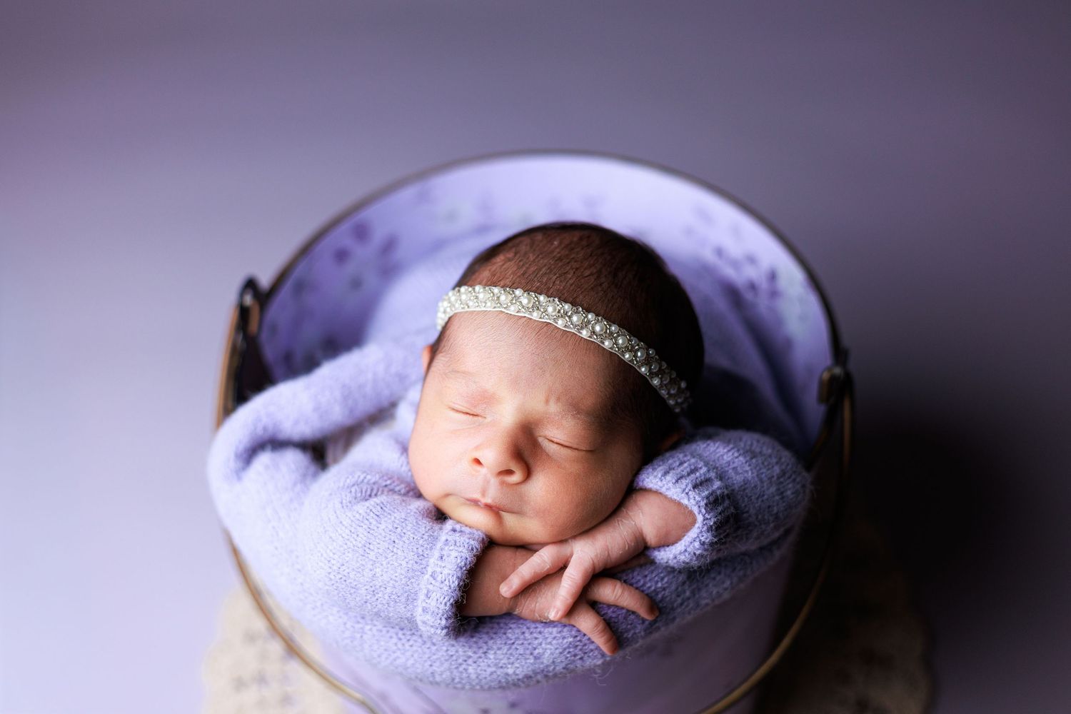 Sleeping newborn baby wrapped in lavender knit blanket with pearl headband, resting in a wooden bucket.