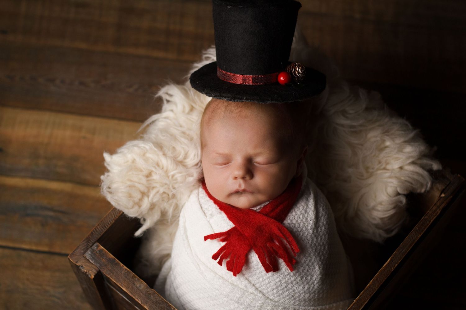 Newborn baby dressed in a snowman-inspired outfit with a black top hat and red scarf during a styled newborn session.