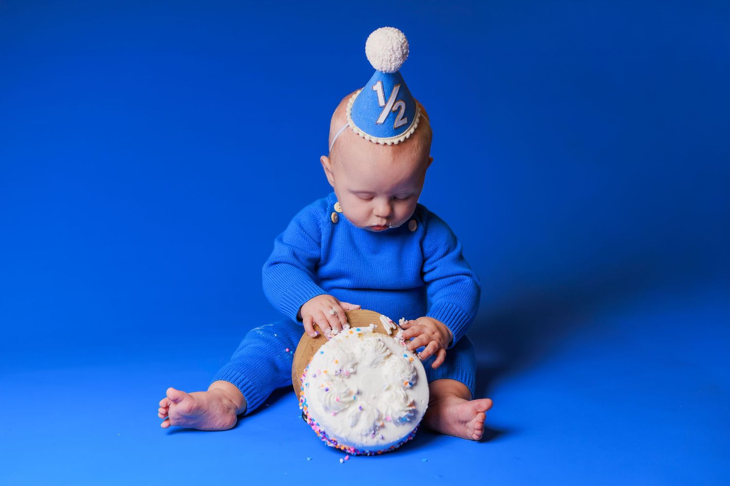 Baby in blue outfit wearing a half birthday crown sits on blue background with a small white cake.