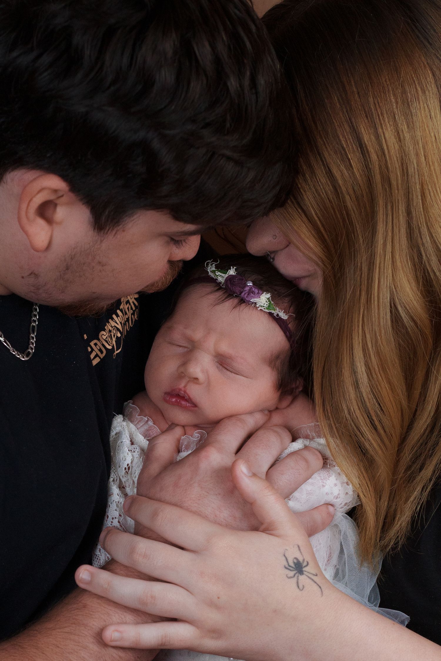 Newborn girl posed with parents snuggled close giving her a kiss on each side.