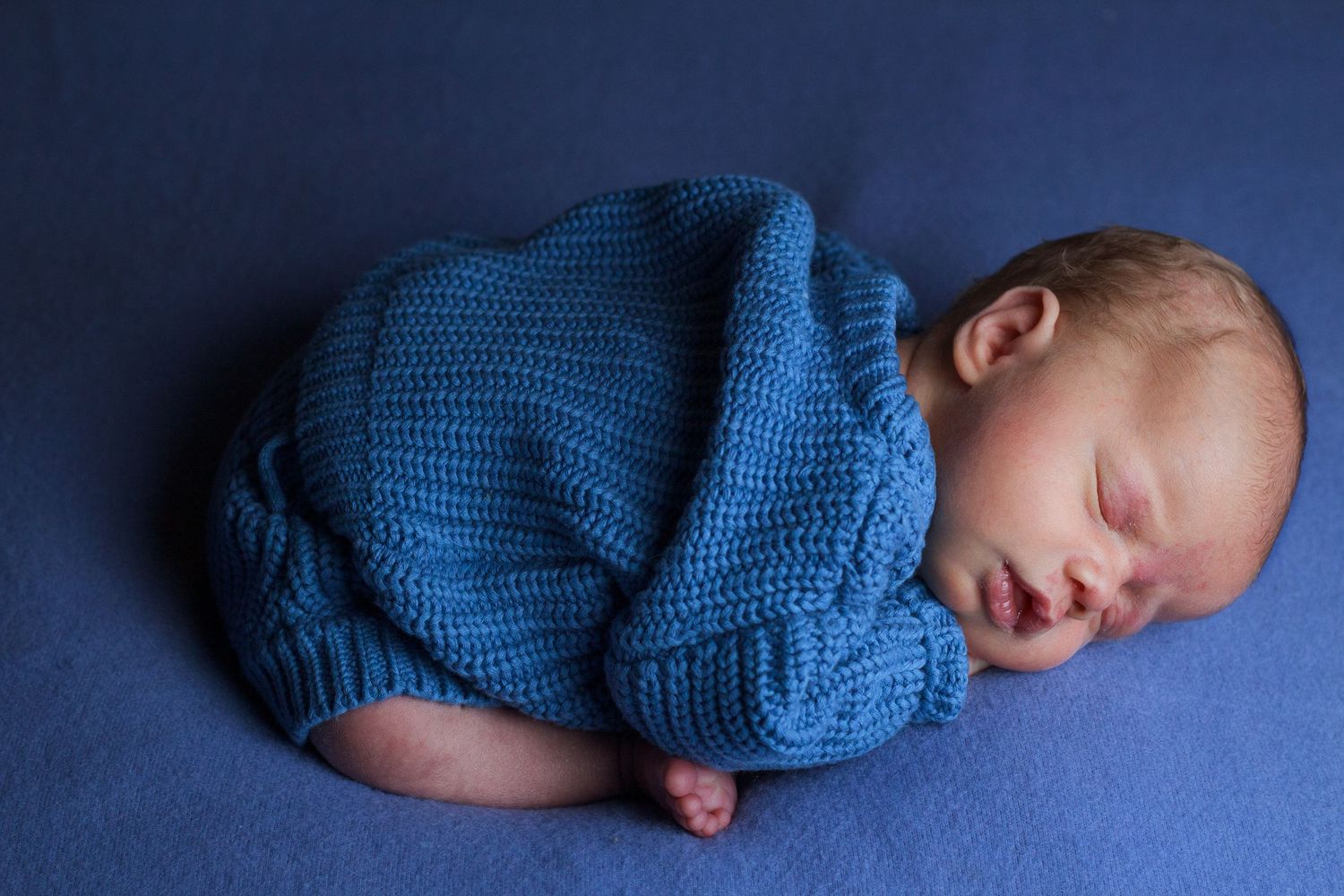 Newborn baby boy sleeping on a soft blue backdrop while wrapped in a textured blue knit.