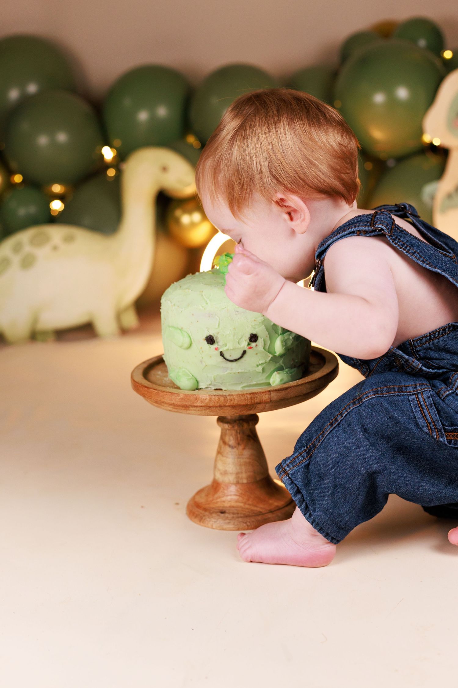 Baby Boy in denim overalls crouched down taking a bite of a dinosaur cake during a cake smash session in Dalton