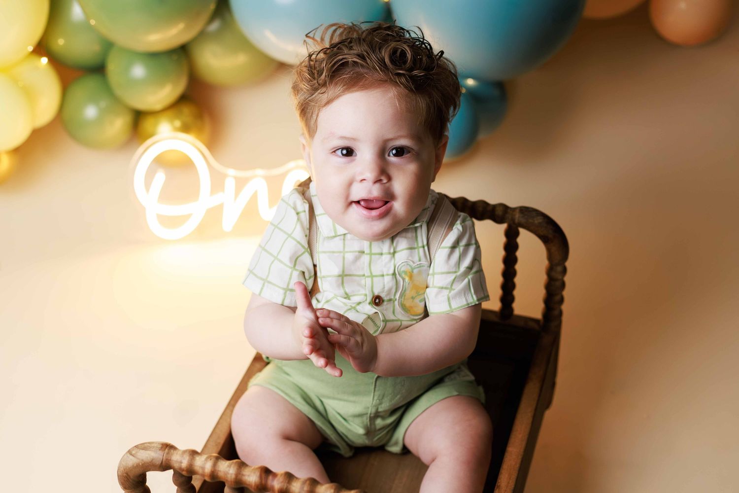 Smiling curly-haired baby boy in plaid shirt at first birthday celebration with colorful balloon backdrop.