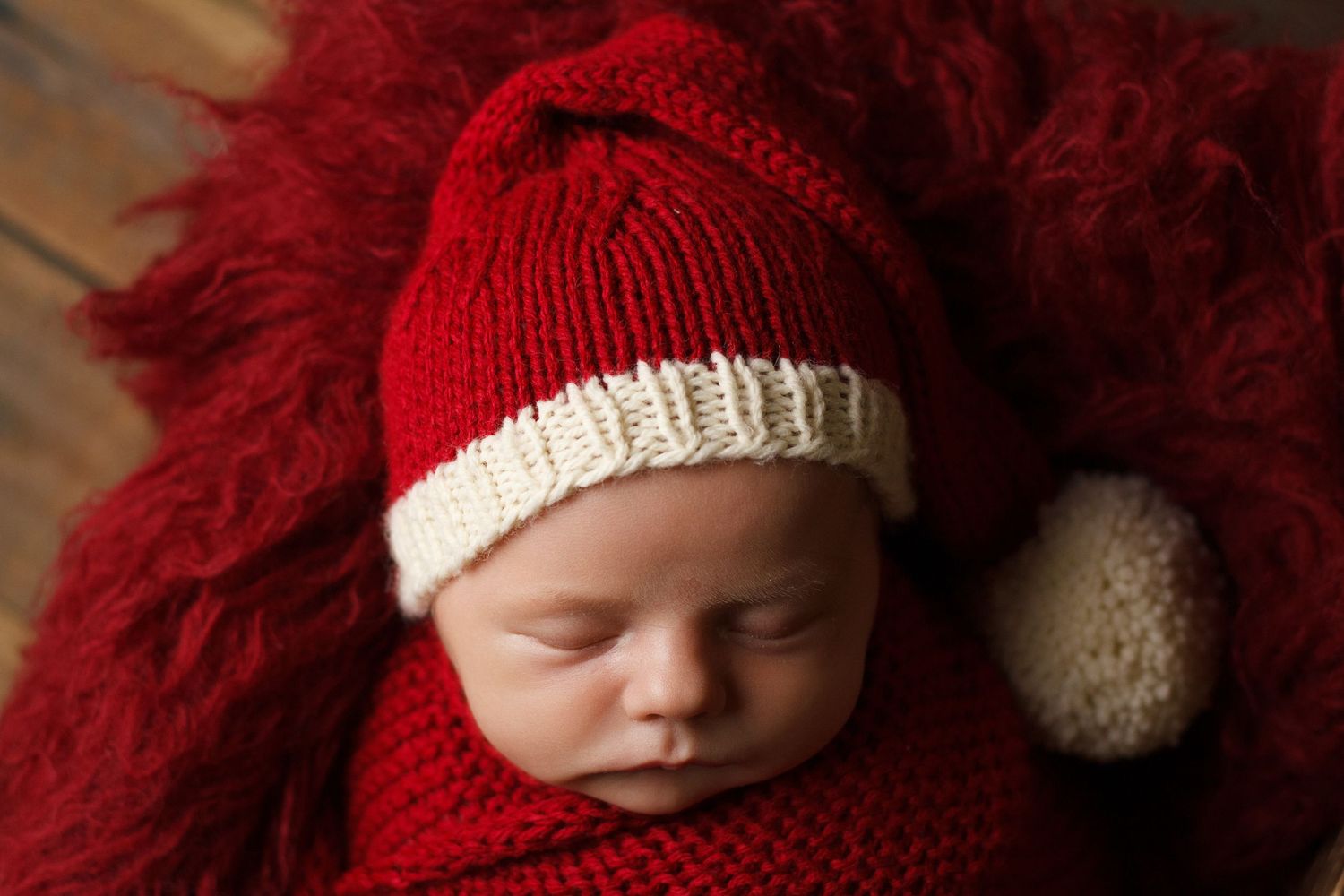 Newborn baby boy sleeping in a red knit hat and wrap on a deep red fur layer.