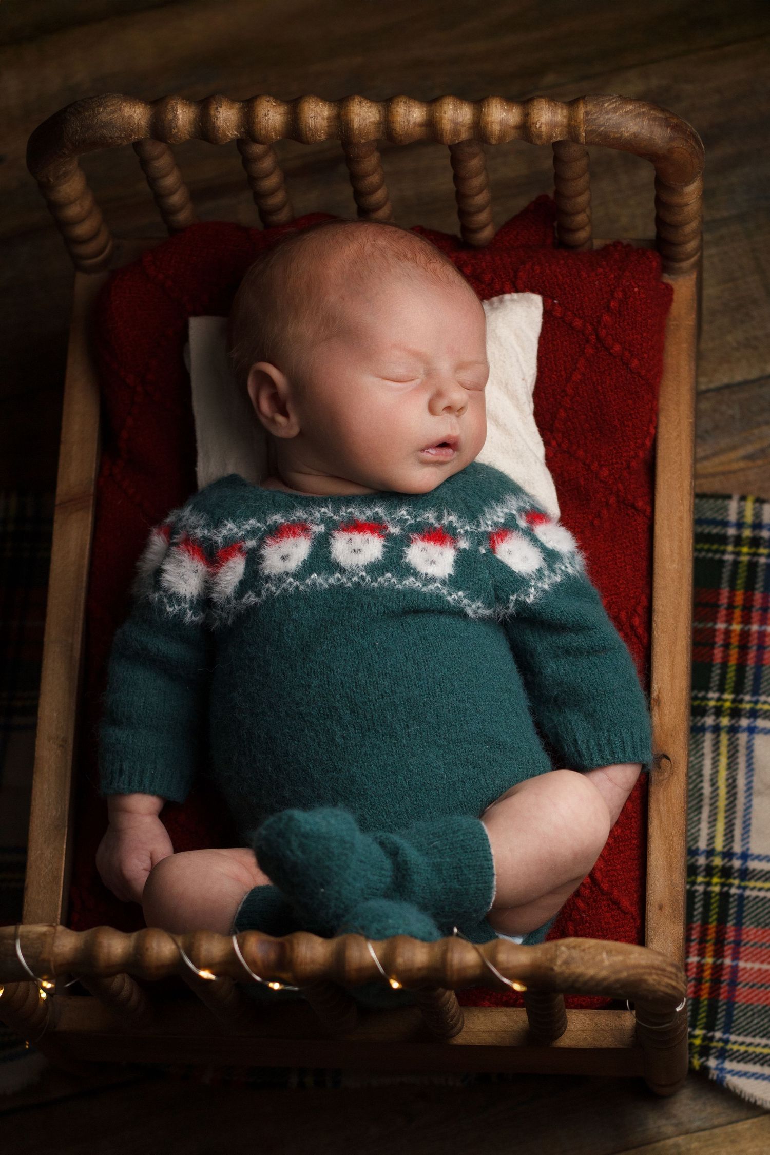 Newborn baby sleeping in a small wooden bed wearing a green knit christmas sweater during a cozy newborn photography session.