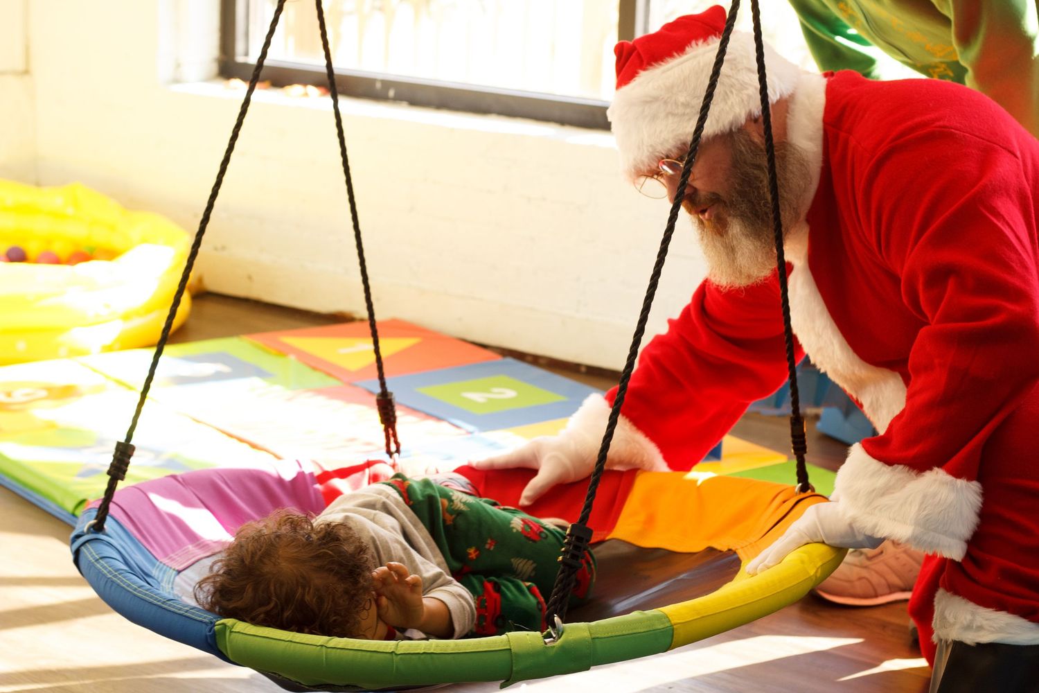 Santa Claus gently interacts with a child lying in a colorful swing indoors.